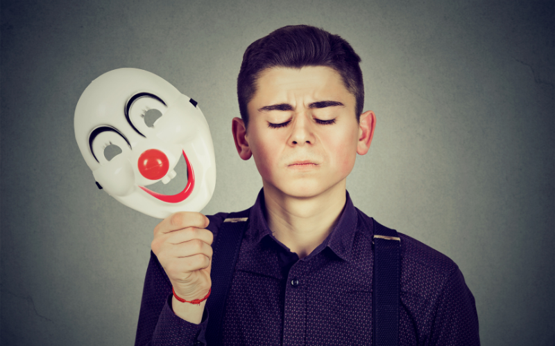 Man frowning while holding a smiley clown face mask — representing the contrast between genuine positivity and forced emotions.